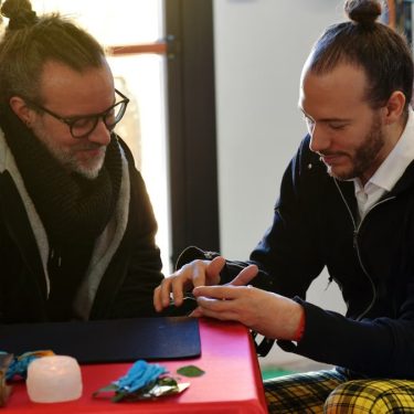 Two men engage in a casual conversation at a table, indoors, with bookshelves in the background.