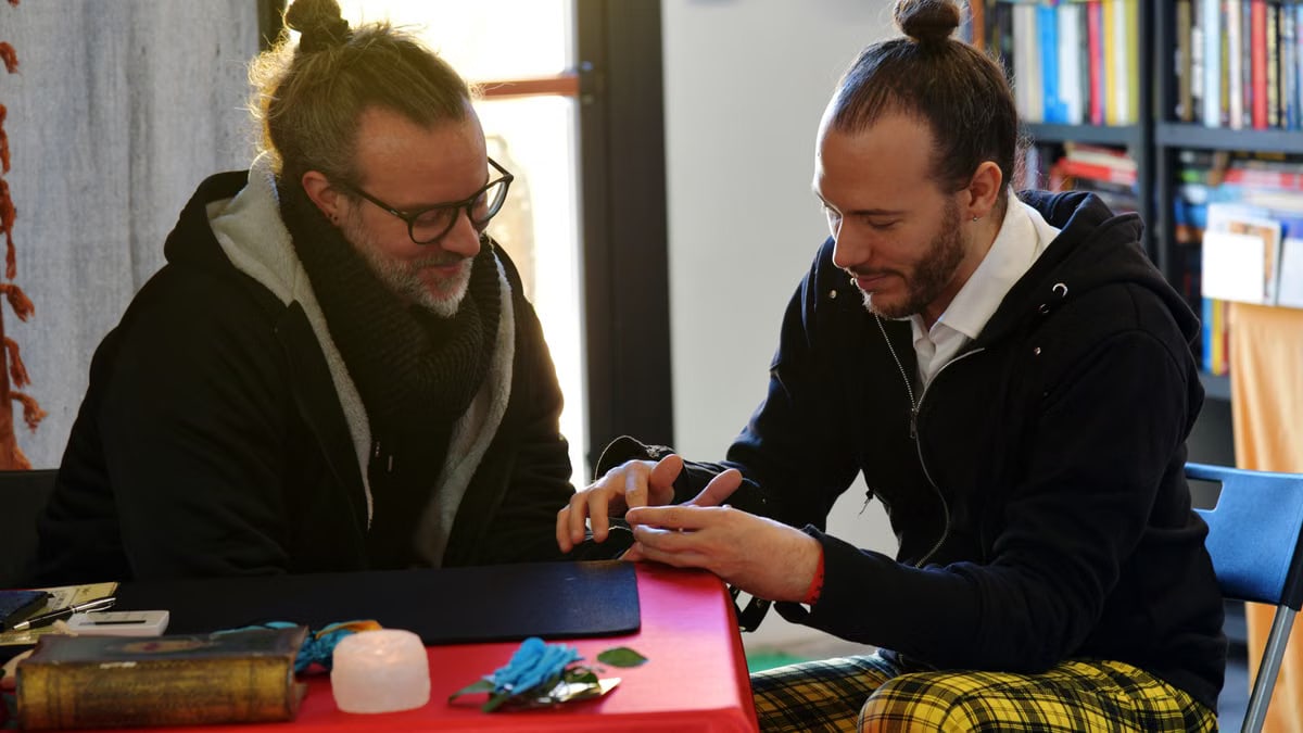 Two men engage in a casual conversation at a table, indoors, with bookshelves in the background.
