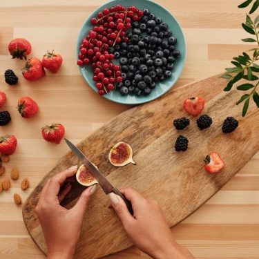 Overhead shot of fresh fruits, nuts, and hands preparing ingredients on a wooden board.